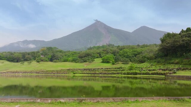 Scenic view of active Fuego Volcano in Guatemala with smoke rising from the peak. Lush green tropical forest surrounds a calm lake with beautiful mountain reflections under hazy sky.