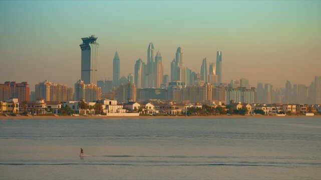 Dubai cityscape viewed across calm waters with lone paddleboarder gliding past luxury villas on Palm Jumeirah. Iconic Marina skyscrapers rise through golden haze as warm sunset light bathes the scene.