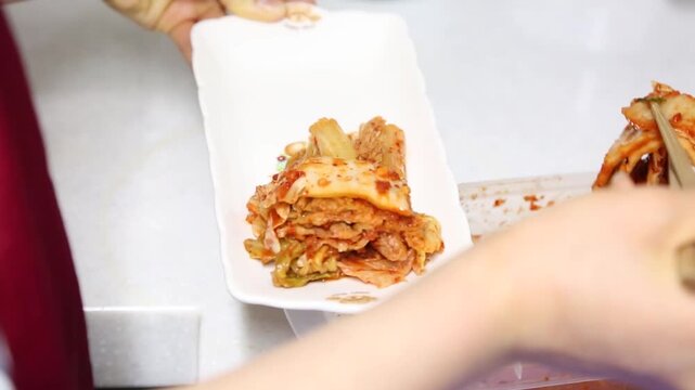 한국 전통 배추김치 접시 상차림 손동작 디테일 자재: 실내 주방에서 젓가락을 이용해 밀폐용기에 담긴 잘 익은 배추김치를 꺼내어 하얀 접시에 정갈하게 옮겨 담는 일상적인 손동작을 포착한 클로즈업 영상입니다. 양념을 담았으며 K-푸드 요리, 한국 가정식 집밥, 전통 발효 식품 및 라이프스타일 콘셉트의 역동적인 인서트 숏입니다.