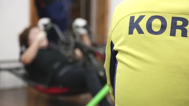실내 훈련장 자전거 코치 KOR 로고 선수 아웃포커스 자재: 실내 훈련장에서 대한민국 KOR 등판 문자가 새겨진 유니폼을 입은 사이클 조력자의 뒷모습을 우측 전경에 타이트하게 배치하고, 그 너머로 페달링하는 장애인 선수를 아웃포커스로 담은 클로즈업 영상입니다. 패럴림픽 자전거 훈련 스케치, 스포츠 팀워크, 지도자 및 한계 극복 콘셉트의 감성적인 인서트 숏을
