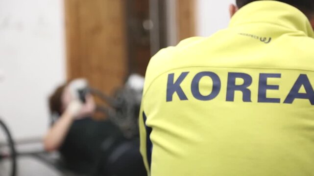 실내 훈련장 자전거 코치 등판 로고 선수 아웃포커스 자재: 실내 훈련장에서 KOREA 로고가 새겨진 상의를 입은 사이클 조력자의 뒷모습을 전경에 배치하고, 그 너머로 훈련 중인 장애인 선수를 아웃포커스로 담은 클로즈업 영상입니다. 팀워크를 담았으며 패럴림픽 자전거 훈련 스케치, 스포츠 지도자, 어시스트 및 한계 극복 콘셉트의 감성적인 인서트 숏을 상징합니다