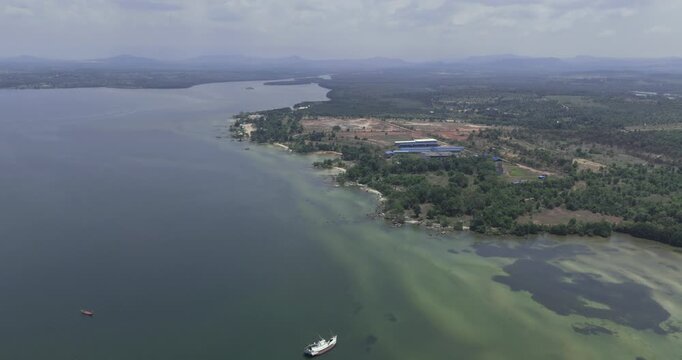 Backward aerial view shows a cargo ship anchored in a calm bay next to a beached fishing boat, with a vast, forested coastline and distant mountains stretching into the haze.