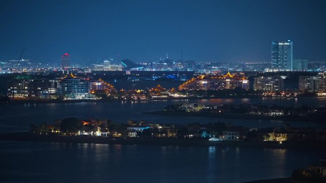 Stunning nighttime aerial view of Dubai waterfront featuring traditional golden-lit Thai-style resort buildings along the bay with modern high-rise towers and city lights glowing against deep blue.