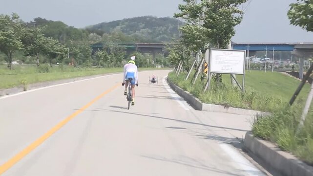 야외 도로 자전거 코치 핸드사이클 추적 주행 자재: 맑은 날 야외 코스에서 일반 자전거를 탄 조력자가 앞서 달리는 삼륜 핸드사이클 선수를 따라가는 모습을 뒤에서 포착한 팔로잉 와이드 숏 영상입니다. 전신주와 고가 교각을 담았으며, 패럴림픽 사이클 훈련, 동반 주행, 팀워크 스케치 및 극복과 도전 콘셉트의 생동감 넘치는 로드 주행 인서트 숏을 상징합니다.