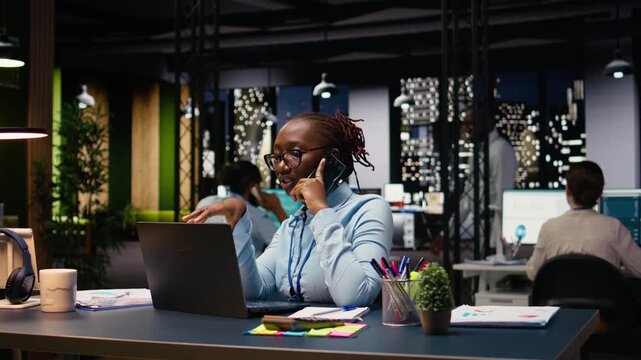 Black employee sharing insights on telephone call in office at night, collaboration between departments to meet objectives and project deadlines. Administrative tasks after hours.