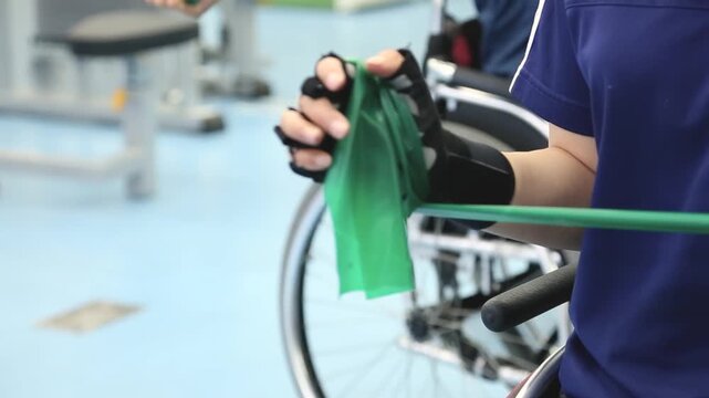 실내 체육관 장애인 휠체어 선수 저항 밴드 근력 운동 자재: 실내 훈련장에서 반장갑을 낀 사용자가 초록색 라텍스 스트레칭 밴드를 손에 감고 당기며 팔 근육 강화 훈련을 하는 모습을 포착한 클로즈업 영상입니다. 배경의 휠체어를 담았으며, 장애인 체육, 저항 운동, 재활 치료, 체력 단련 및 극복과 도전 콘셉트의 실감 나는 인서트 숏을 상징합니다.