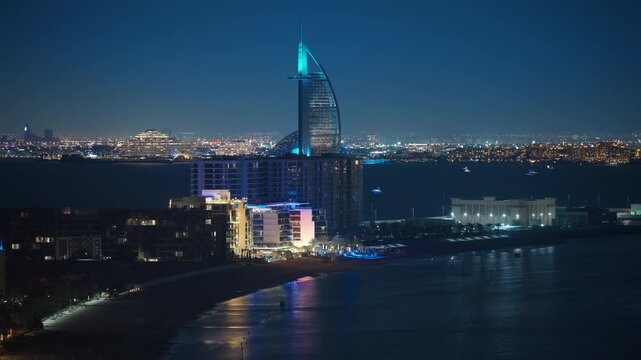 Stunning nighttime aerial view of Dubai coastline featuring the iconic sail-shaped tower illuminated in turquoise blue, surrounded by luxury hotels along the sandy beach with city lights sparkling.