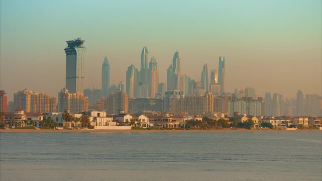 Stunning Dubai Marina skyline viewed across calm waters during golden hour with luxury Palm Jumeirah beachfront villas in foreground, hazy atmospheric conditions softening the towering skyscrapers.