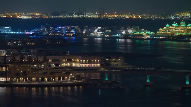 Dubai coastal development featuring luxury waterfront resorts and residential villas illuminated at night. Colorful lights reflect on calm waters with industrial port glowing in distant background.