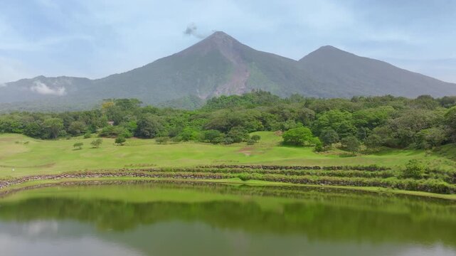 Aerial drone footage panning across a scenic golf course with calm reflective pond and stone terraces, revealing majestic smoking volcano peaks rising above dense tropical rainforest under hazy skies.