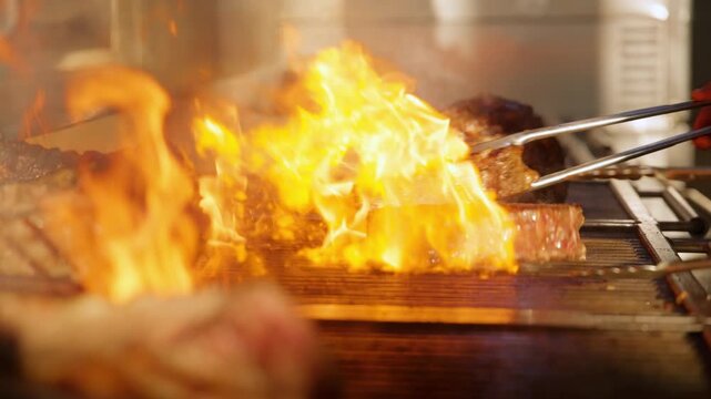 Chef uses metal tongs to flip thick premium beef steaks on a commercial grill as bright orange flames flare up dramatically, creating beautiful searing marks in a professional restaurant kitchen.