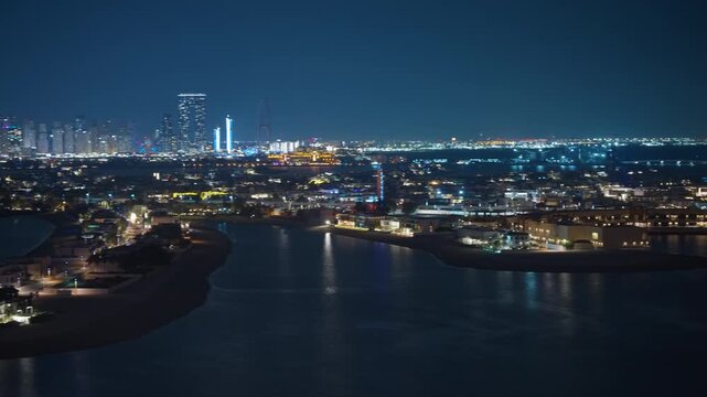 Aerial drone footage captures Dubai skyline illuminated at night with glowing skyscrapers of Dubai Marina in background. Luxury waterfront villas on Palm Jumeirah fronds reflect in calm waters below.