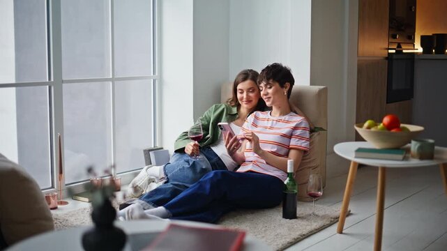 Smiling roommates looking smartphone relaxing together in modern apartment.