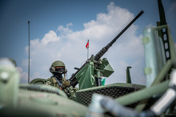 Close-up of a military soldier wearing tactical gear and a helmet operating a heavy machine gun mounted on a turret during an army exercise. © Sirirat Makprasert