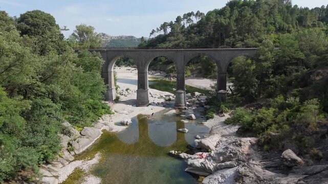 Cinematic drone shot flying under a historic stone arch bridge over the Gardon river. Rising up to reveal the beautiful valley and the famous Porte des Cevennes rock formation near Anduze, France.