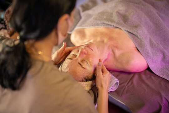Professional dermatologist making relaxing beauty facial massage for woman lying on table under towel, female patient enjoying lifting drainage massage session, self love and feminine beauty rituals.