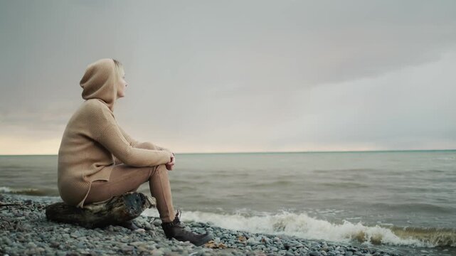 Young woman sitting on wooden log on pebble beach looking at ocean waves under cloudy sky.