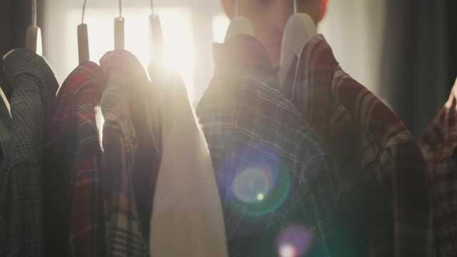 Young man choosing clothing from rack in bright sunlit room with casual shirts hanging on hangers inside wardrobe close up shot.