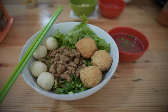 A bowl of chicken noodles with fish balls and green vegetables. Green chopsticks are placed beside the dish. A bowl of soup is nearby.