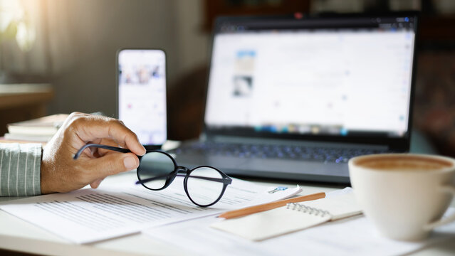 close up shot, man hand holding eyeglasses working online at home with business documents and laptop start new business, E-commerce