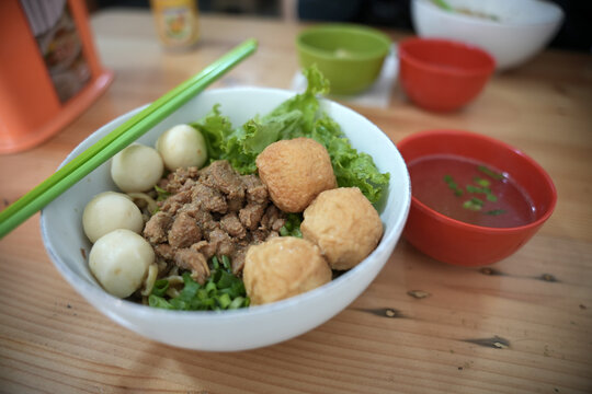 A bowl of chicken noodles with fish balls and green vegetables. Green chopsticks are placed beside the dish. A bowl of soup is nearby.