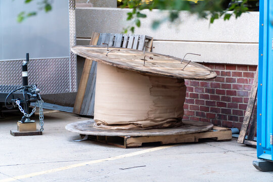 Large weathered wooden cable spool wrapped in brown canvas, standing on concrete at a bustling construction site with a trailer hitch, brick wall, and blue portable toilet in an urban setting.