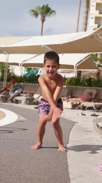 Front view of playful child dancing barefoot near swimming pool at family resort on sunny summer day. Vertical reel of cheerful male kid having rest at water pool on summer vacation.