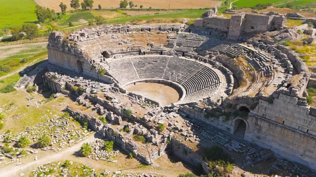 Balat, Turkey. The ancient Theatre of Miletus, its semicircular stone cavea and ruined stage building rising from the archaeological site, aerial view over green farmland and distant mountains. Aeria