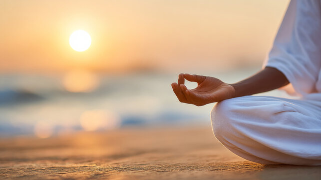 A person in white clothing sits in a meditation pose on a beach at sunset, with a focus on their hand in a mudra gesture against the softly blurred ocean horizon.