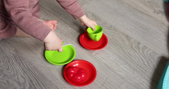 Little child playing with colorful toy tableware set sitting on floor. Toddler girl hands holding green plastic teacup and white fork above bright plates. Educational sensory roleplay game development