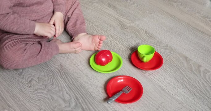 Little child sitting on floor playing with colorful plastic toy tableware kitchen set. Toddler hands holding white fork picking red toy tomato on green plate. Educational roleplay game preschool