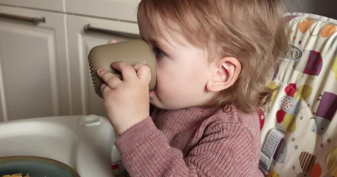 Mother hand helping little child drink from silicone cup sitting in highchair. Cute toddler girl holding mug drinking water milk juice kitchen interior background. Care complementary feeding process