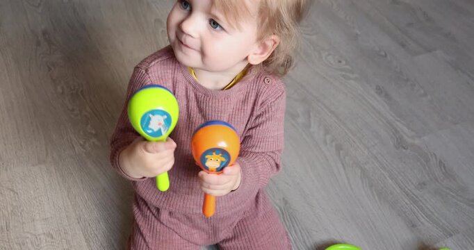 Cute little child holding colorful maracas musical instruments sitting on floor. Happy toddler girl shaking bright plastic rattles making music indoor home background. Early childhood development