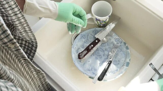 Man in green rubber gloves washes dishes in a kitchen sink using a sponge and liquid soap. Contemporary domestic scene and modern masculinity. Concept of sharing household chores. Day light, close-up