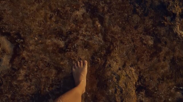 A first-person view of a person walking barefoot through calm, crystal-clear shallow seawater over a rocky, sandy seafloor, capturing a serene summer beach vacation atmosphere.