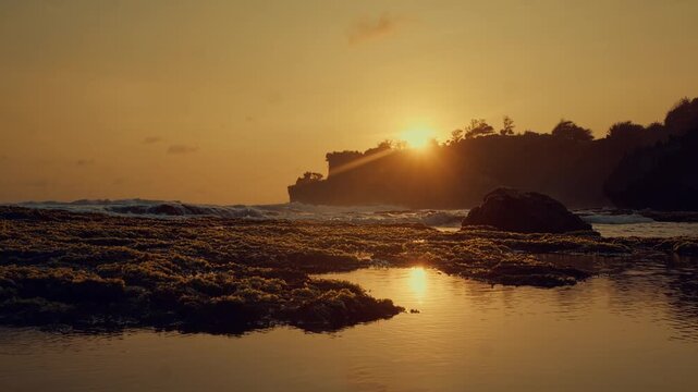 Golden hour sunlight illuminates a rocky beach with tide pools and crashing waves, creating a peaceful and serene coastal atmosphere during a vibrant Bali landscape sunset.