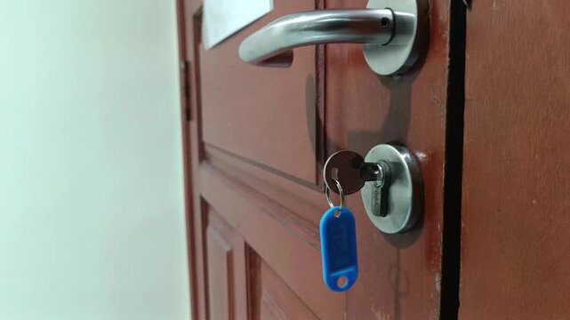 Footage of a metal key with a blue plastic key chain tag still stuck in the keyhole cylinder of a brown wooden door.