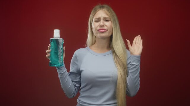 Woman young blonde holding mouthwash bottle with eyes closed and grimacing, right hand raised in studio; disgust aversion oralcare.