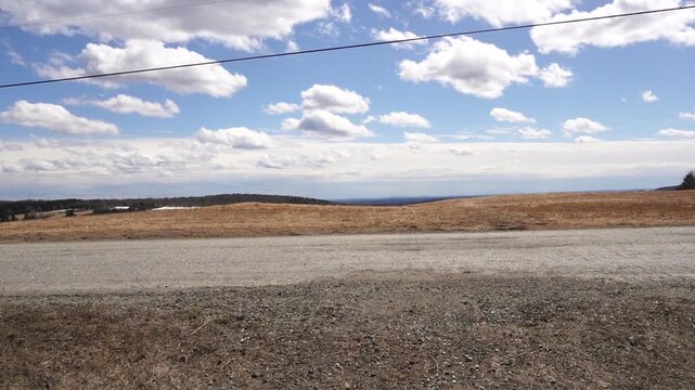 Eye-level, panoramic view of a gravel road bordering a vast, dormant brown agricultural field. Distant rolling hills under a vibrant blue sky with scattered white cumulus clouds. Power line across top
