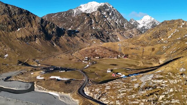 Revealing Aerial View of Winding Coastal Road and Village Surrounded by Snowy Mountains in Haukland Beach at sunset, Lofoten, Norway, 4K 60fs pull away horizontal drone shot