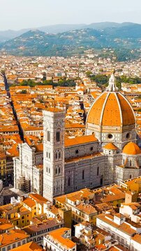 Vertical video. Florence, Italy. Cathedral Santa Maria del Fiore, its vast terracotta-tiled dome and white marble facade rising above the dense historic cityscape, aerial panoramic view under clear s