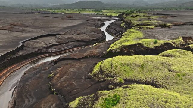 River cutting through ancient volcanic lava fields from Mount Yasur volcano near Port Resolution creates a dramatic and eroded landscape with green vegetation on its banks. Aerial view drone flight