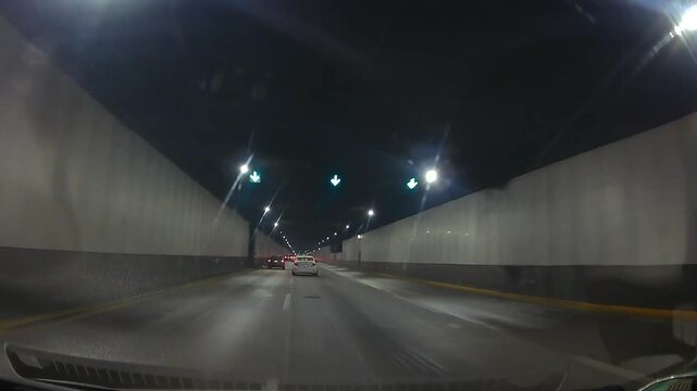 Dashcam POV of night driving through an illuminated urban tunnel with cars, concrete walls, road lights and moderate traffic.