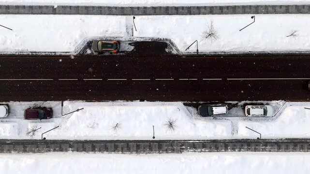 Aerial view shows snow-covered highway. Dark asphalt road stretches through the scene. Bus travels on the road. Cars park beside it on snow. No people present. Quiet winter setting
