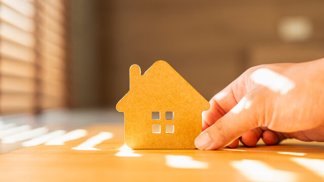Hand touching a wooden house model on a table with warm sunlight. Concept of real estate investment, buying a new home, property insurance, and family future planning.