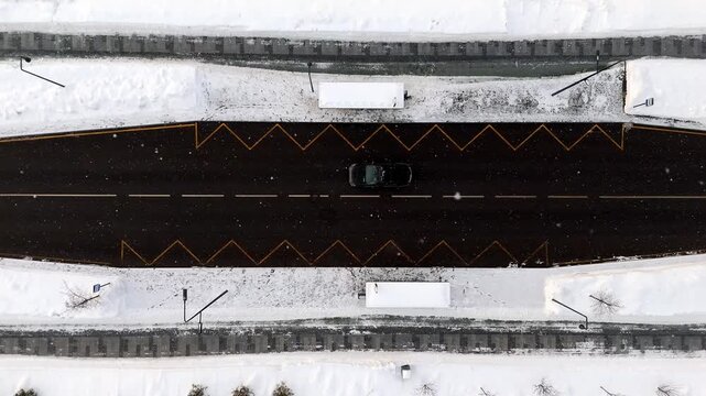Aerial view shows a snow-covered road with yellow zigzag markings. Car travels on the black asphalt. Snow covers the surrounding landscape and trees. The scene highlights winter road conditions