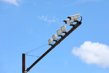 Obraz premium Security cameras and floodlights installed on a high pole under a bright blue sky, representing surveillance technology, public safety, monitoring systems, and urban security infrastructure.