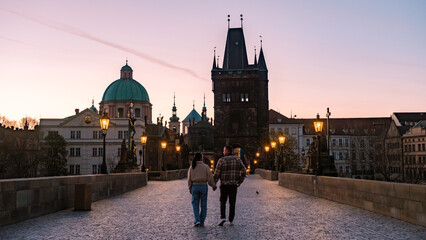 Obraz premium Couple walks hand in hand along the historic Charles Bridge in Prague as the sun sets, casting a magical glow over the city. Lanterns illuminate the cobblestone path, creating a romantic atmosphere.