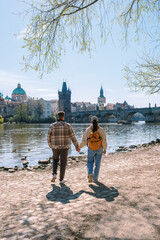 Obraz premium A young couple strolls along the Vltava River, enjoying the beautiful scenery of Prague. They are surrounded by historic buildings and the serene water under a clear blue sky.