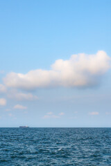 Fototapeta premium Minimalist vertical view of vast blue sky with giant fluffy white clouds over calm ocean water and a tiny distant cargo ship on the horizon, perfect copy space.